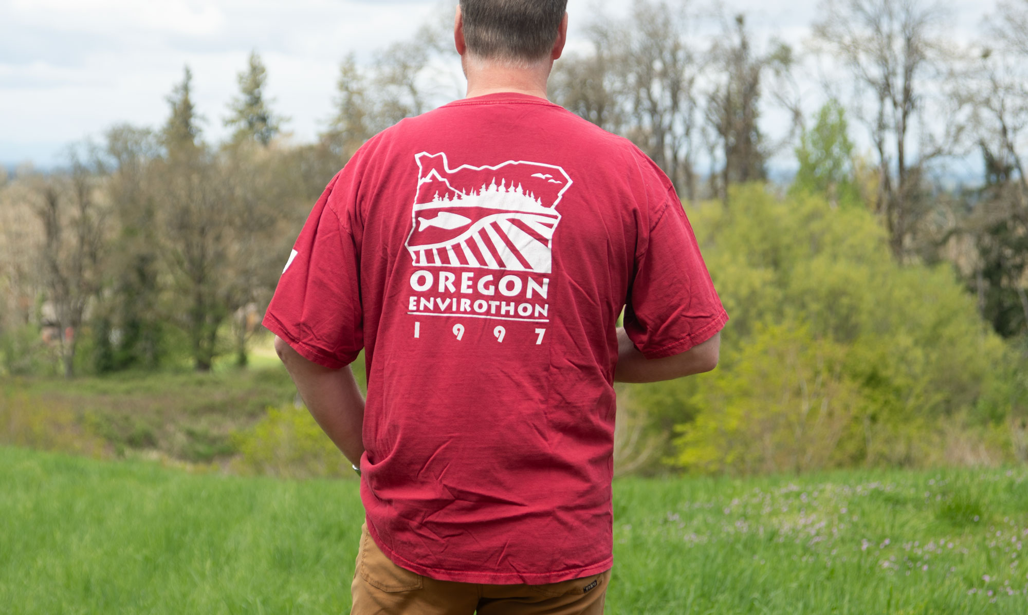 Man standing in a grassy field wearing a red Oregon Envirothon 1997 shirt, shown from the back.