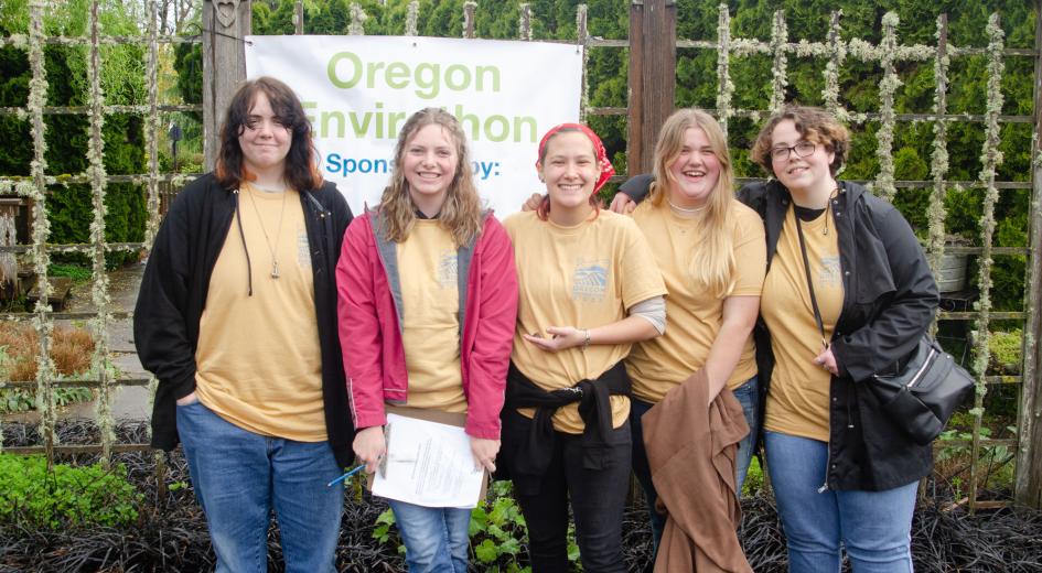 A group of five students in matching Oregon Envirothon shirts pose and smile in front of a banner at the event.