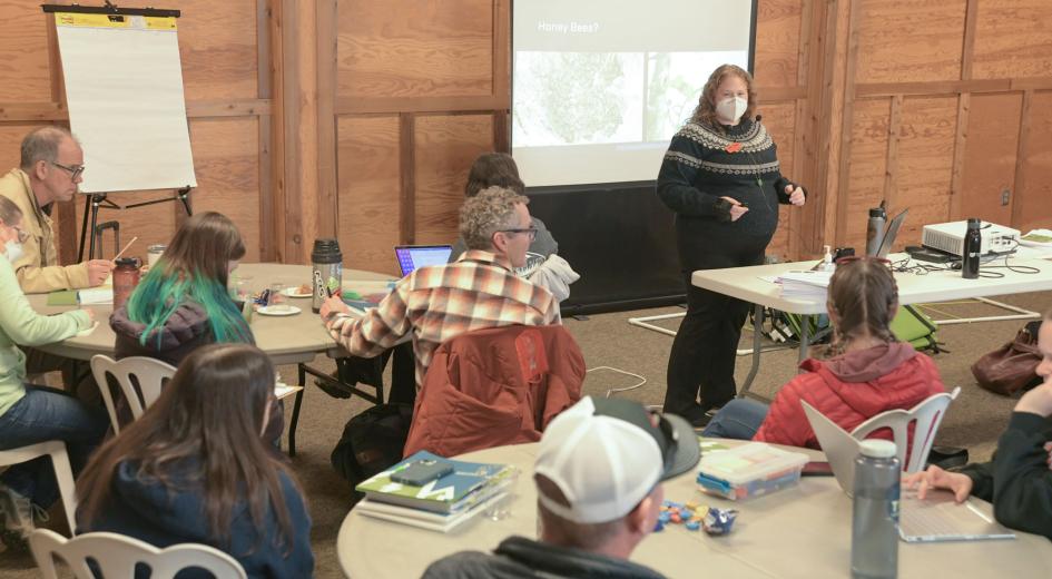 An instructor presents to a group of adults seated at round tables during an indoor educational training session.